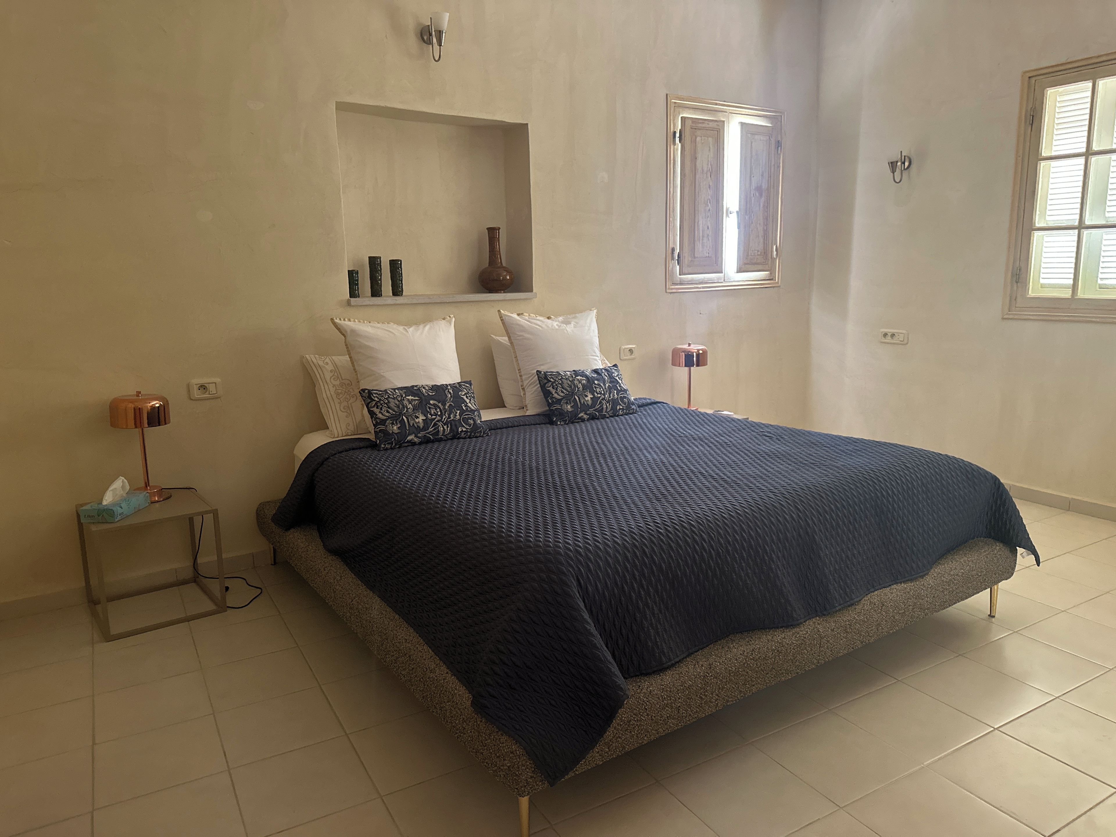 A bedroom with a bed, bedside lamps, a nightstand, a window, and a wall-mounted shelf.