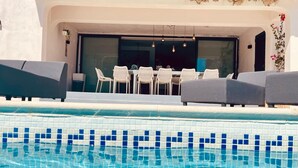 Une piscine avec un bord carrelé, un espace repas comprenant des chaises et une table, et un bâtiment à la façade blanche.