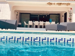 En swimmingpool med flisekant, et spiseområde med stole og et bord, samt en bygning med en hvid facade.