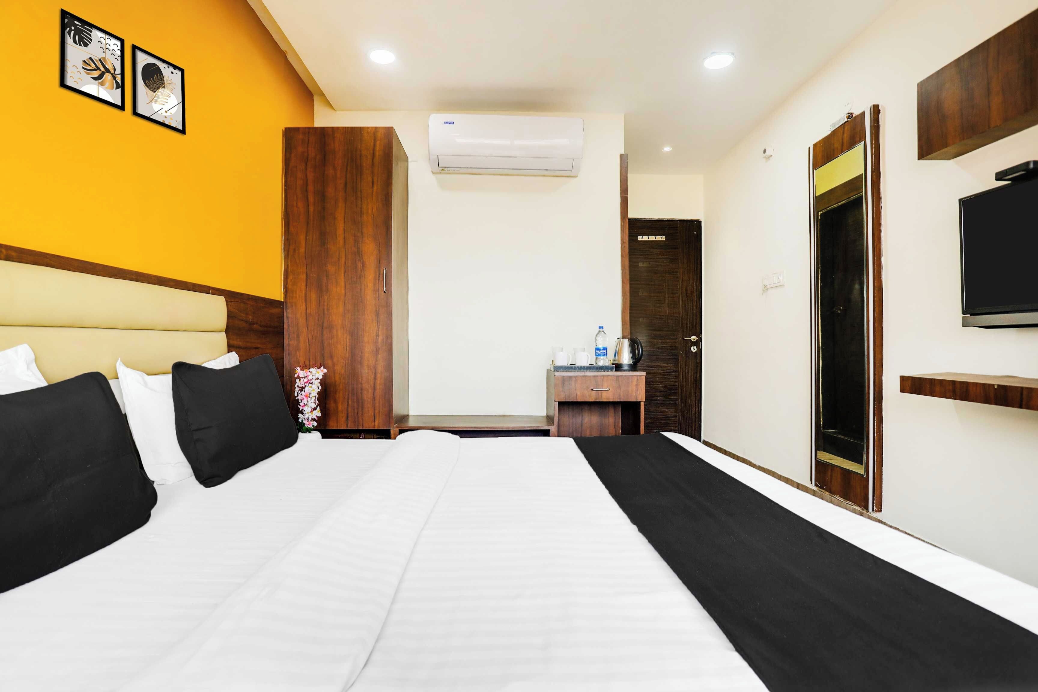 A hotel room with a large bed, a wooden wardrobe, a desk with a kettle, and a television on the wall.