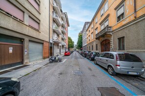 A narrow urban street lined with parked cars and multi-story buildings with balconies. - An elegant, modern 70-square-meter apartment located on the fifth floor of a building (with an elevator, though not suitable for people with disabilities) near the city center.Ideal for two people, the property is situated just a short walk from the trai (Udine)