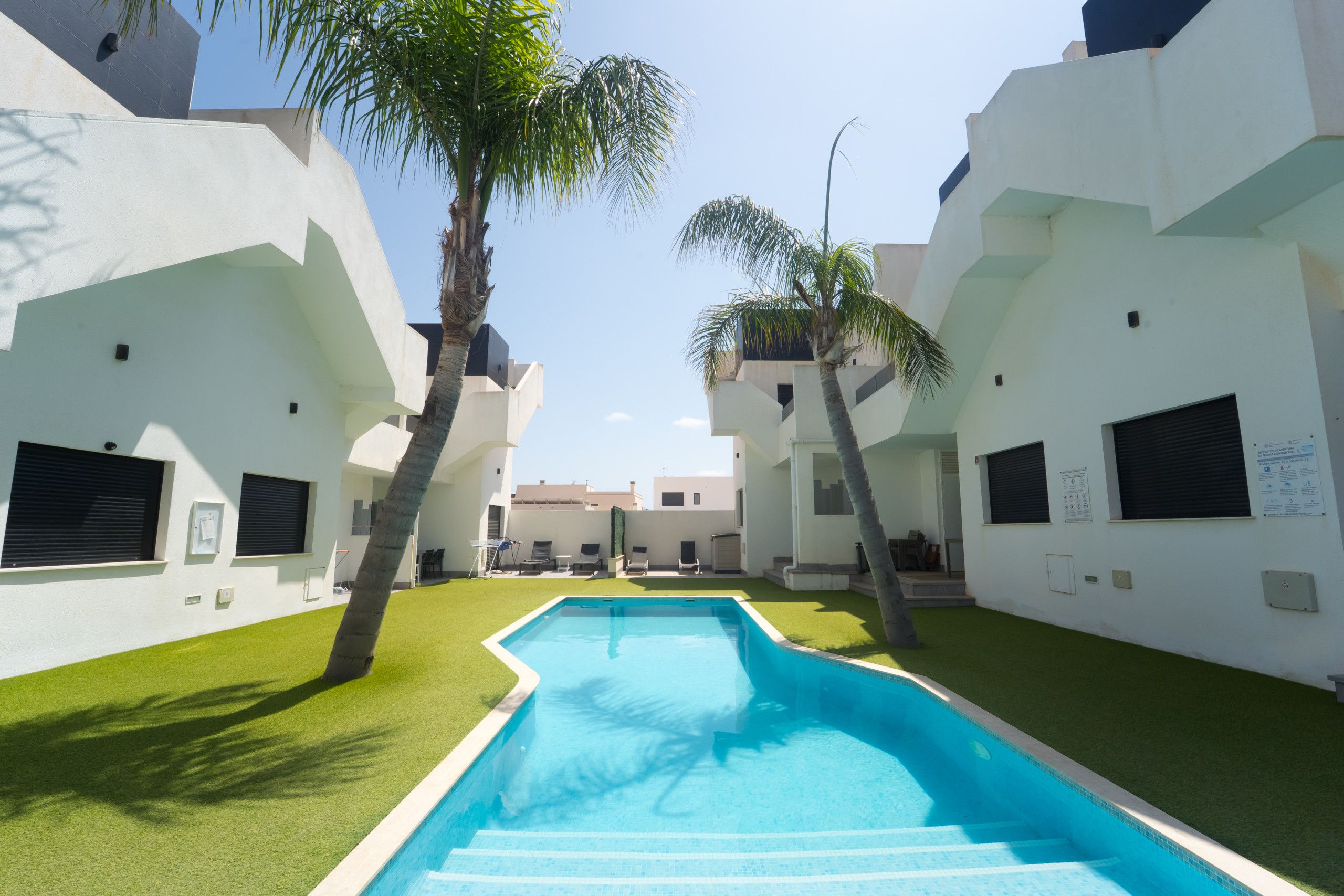 A pool area with a rectangular pool, surrounded by white walls and green artificial grass.
