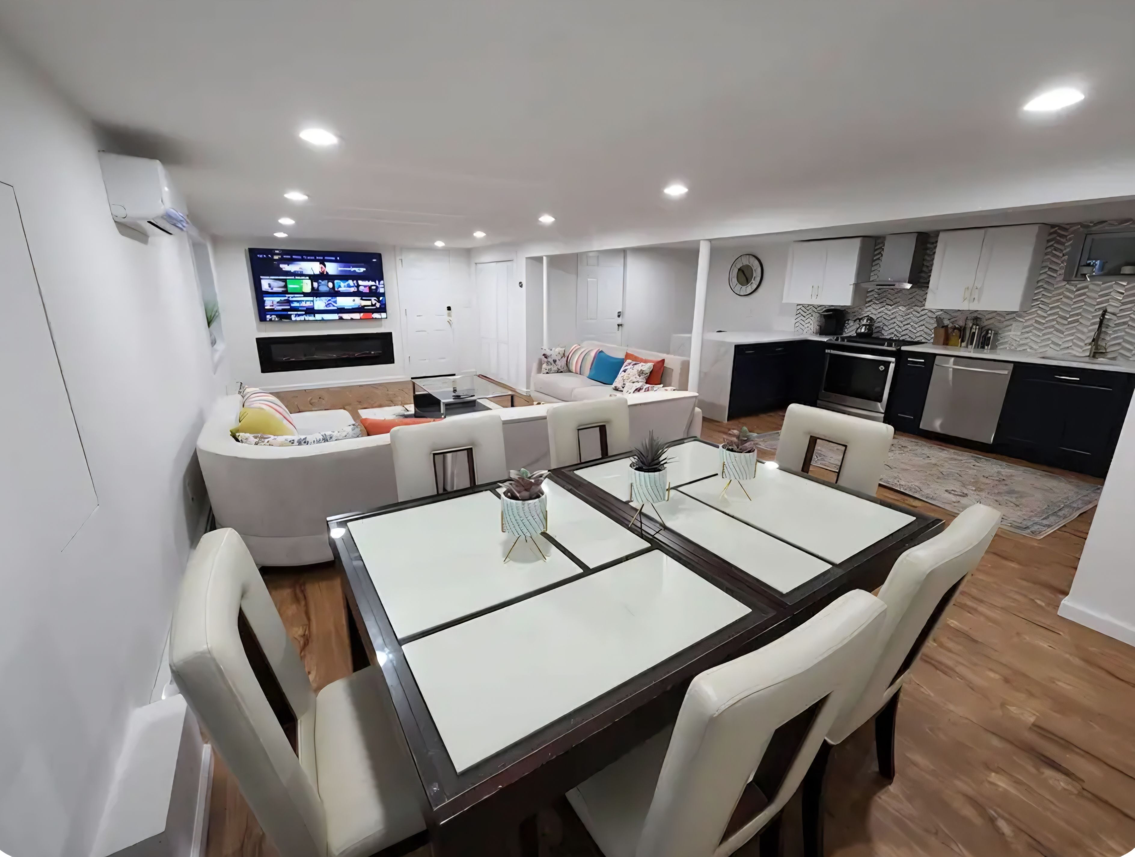 A modern living room with a flat-screen TV, a dining table with chairs, a sofa, and a kitchen area with cabinets and appliances.