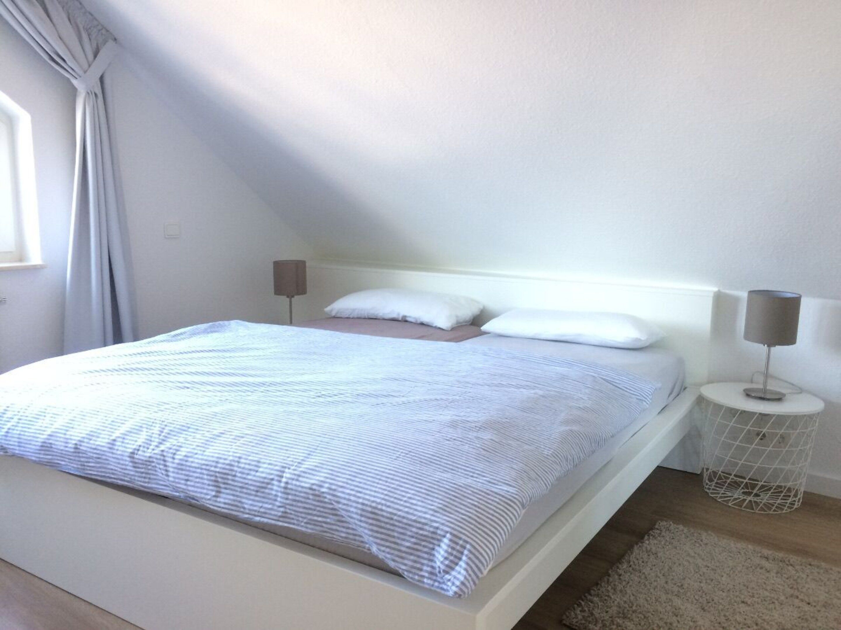 A bedroom with a bed, two bedside lamps, a window with curtains, and a small white rug.