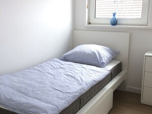 Une petite chambre avec un lit simple, une tête de lit blanche, une fenêtre avec des stores et une petite armoire blanche.