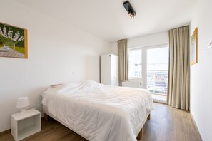 A bedroom with a bed, a bedside table, a wardrobe, a window with curtains, and a painting on the wall. - Apartment in Blankenberge With Sea Views (Blankenberge)