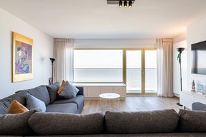 Ein modernes Wohnzimmer mit einem grauen Sofa, einem kleinen runden Tisch und einem großen Fenster mit Meerblick.