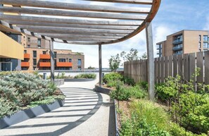 A covered outdoor area with a curved wooden structure, surrounded by greenery and adjacent to buildings with balconies. - Luxury Heathrow Apartment, 20min from London Central (Hounslow)