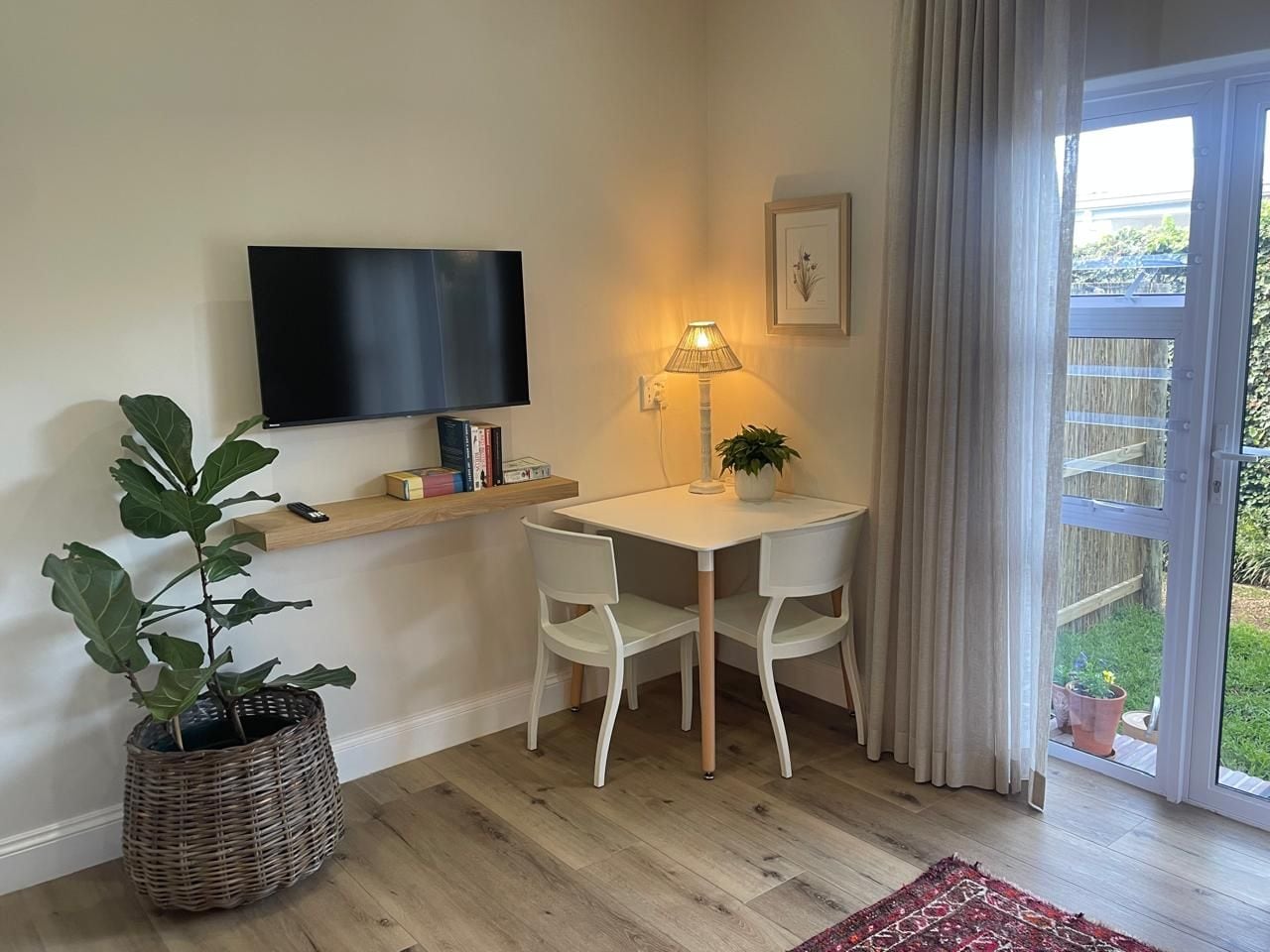 A room with a television mounted on the wall, a table with two chairs, a potted plant, and a sliding glass door leading to an outdoor area.