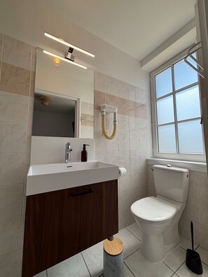 Ein modernes Badezimmer mit Waschbecken, Spiegel und Toilette. Ein Fenster lässt natürliches Licht herein und eine Dusche ist sichtbar.
