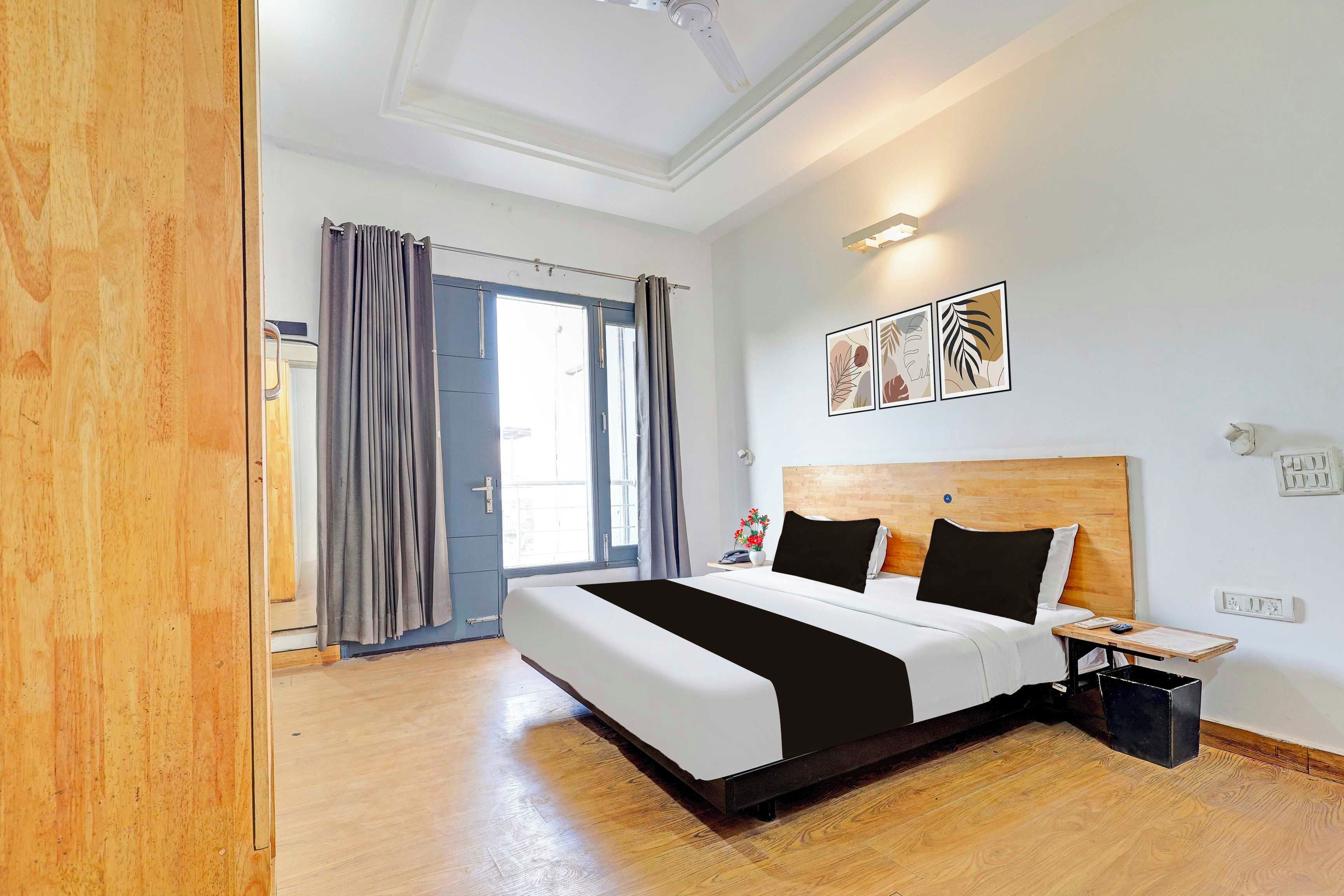 A hotel room with a large bed, a wooden headboard, a bedside table with a lamp, and a window with curtains.