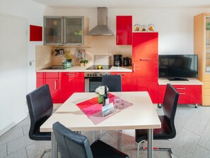 Eine kompakte Küche mit roten Schränken, einem Esstisch mit Stühlen, einem Herd und einem Fernseher auf einem Schrank.