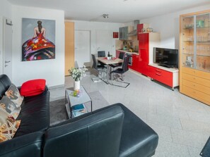 Ein modernes Wohnzimmer mit einer schwarzen Couch, einem Essbereich mit Tisch und Stühlen sowie einer Küche mit roten Schränken und einem Fernseher.