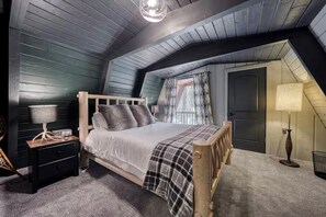A bedroom with a sloped ceiling, a bed with a wooden frame, a nightstand with a lamp, and a window with curtains. - Modern A-Frame Cabin with Hot Tub Near Yellowstone, Island Park, Idaho (Island Park)