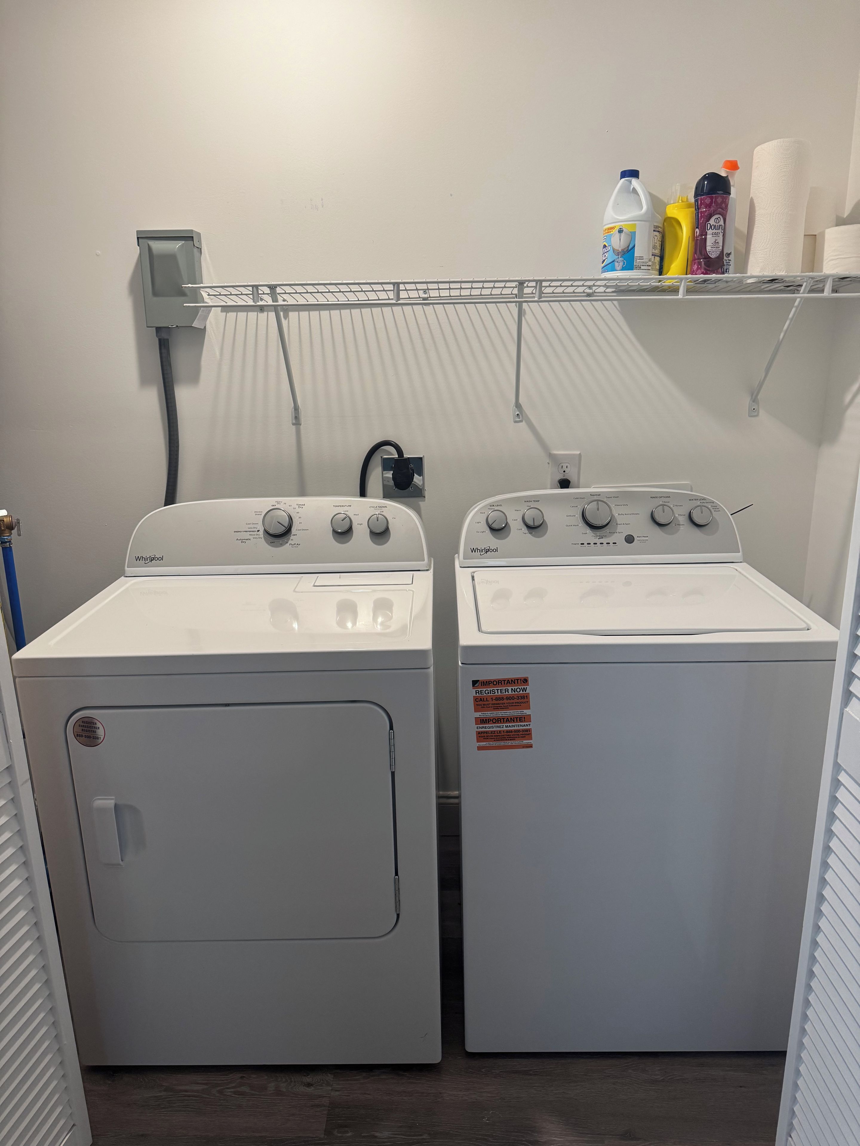 A laundry room with two white Whirlpool washers, a power outlet, and a shelf with cleaning supplies.