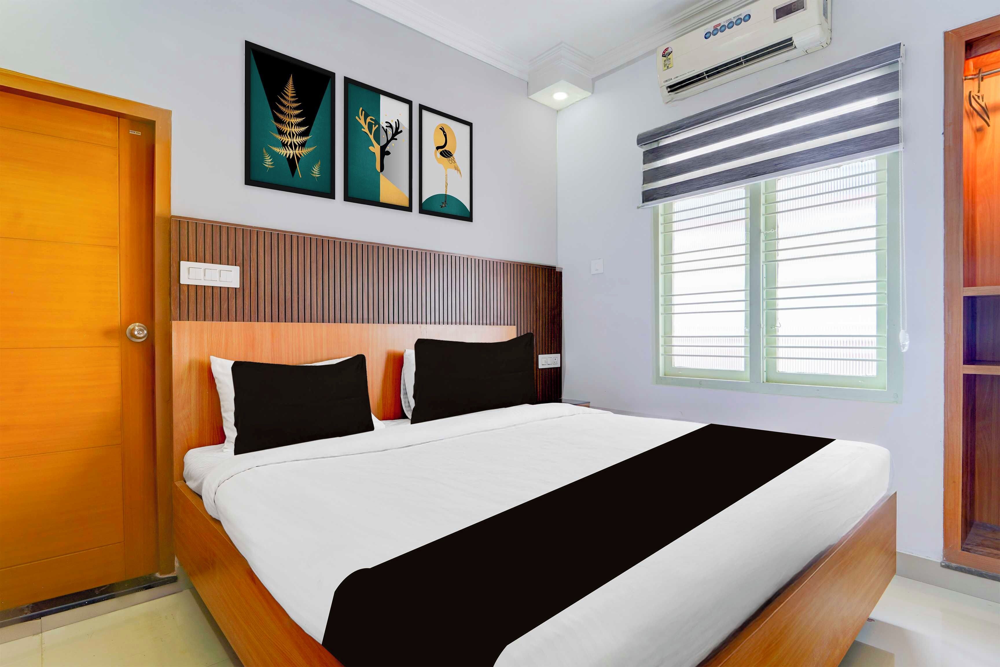 A hotel room with a bed, a wooden headboard, a window with blinds, and three framed artworks on the wall.