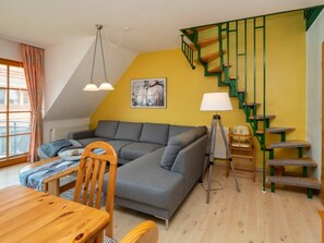 A cozy living room with a grey sofa, wooden dining table, and a green staircase. - 1.5/t3 Holiday Home Park \"freesenbruch\" (Zingst)