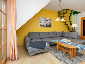 A living room with a grey sofa, a wooden coffee table with magazines, a staircase with green railings, and a painting on the wall. - 1.5/t3 Holiday Home Park \"freesenbruch\" (Zingst)