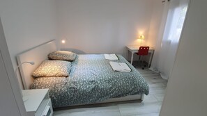 Ein ordentlich bezogenes Bett mit gemustertem Überwurf und Kissen, ein Nachttisch mit Lampe sowie ein kleiner Schreibtisch mit Stuhl.