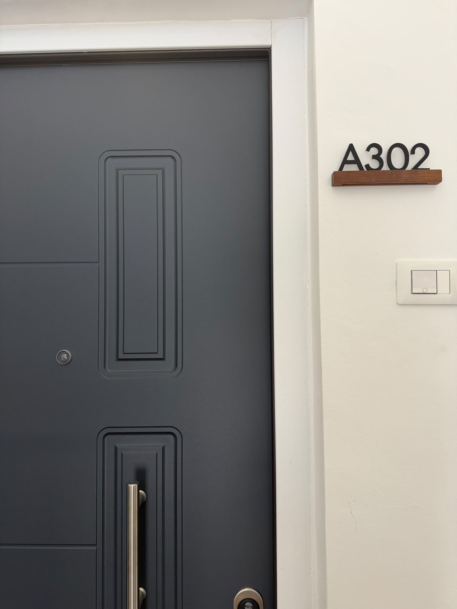 Une porte gris foncé, dotée d’une poignée et d’un trou de serrure, un mur blanc sur lequel est fixée une plaque portant le numéro « A302 », et un interrupteur.