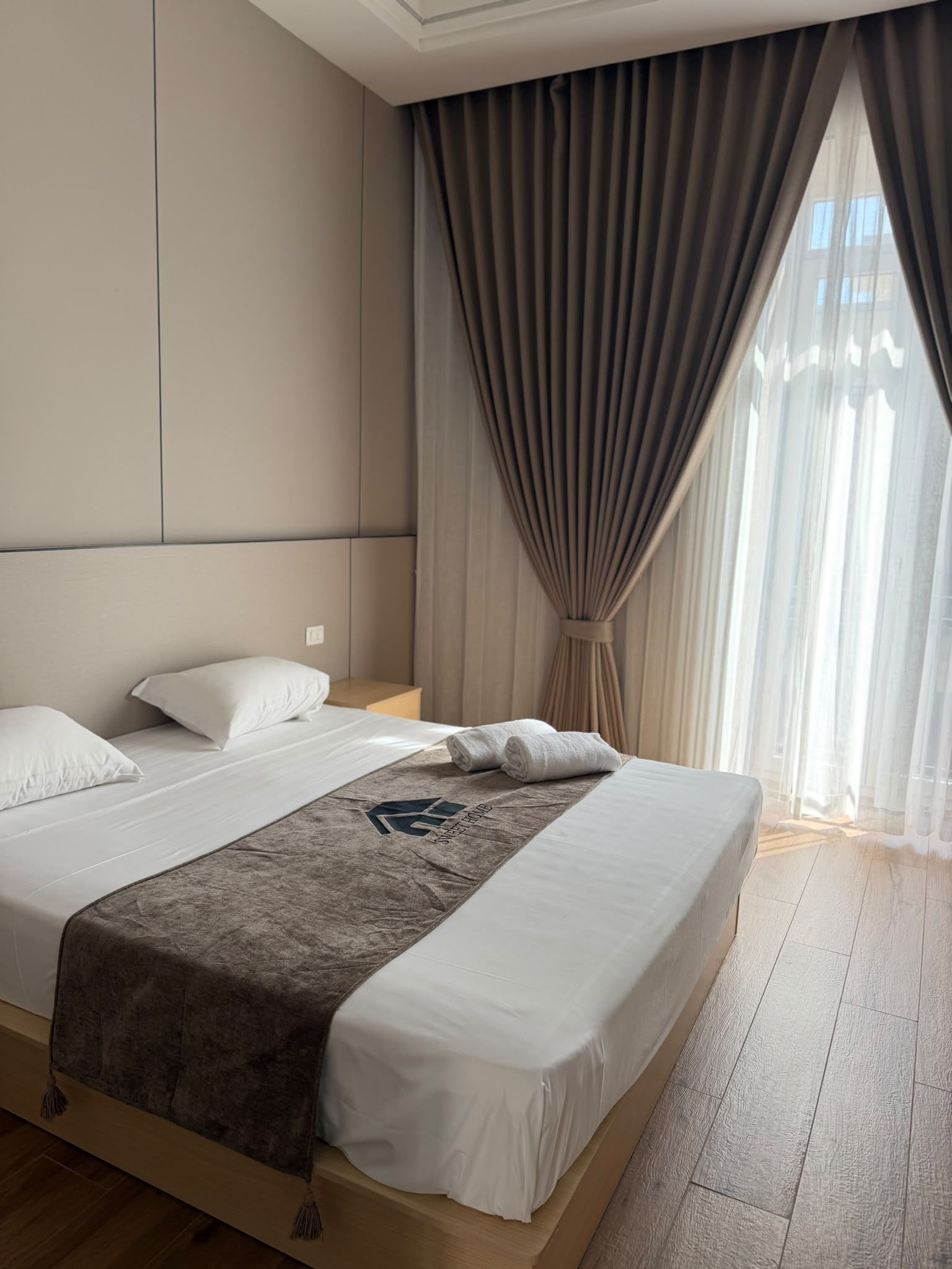 Une chambre d’hôtel avec un grand lit, une tête de lit, une table de chevet et une fenêtre avec des rideaux.