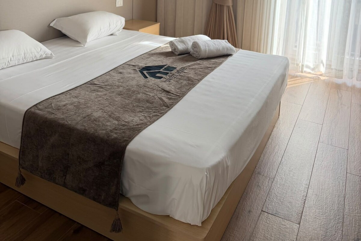A hotel room with a large bed, a headboard, a nightstand, and a window with curtains.