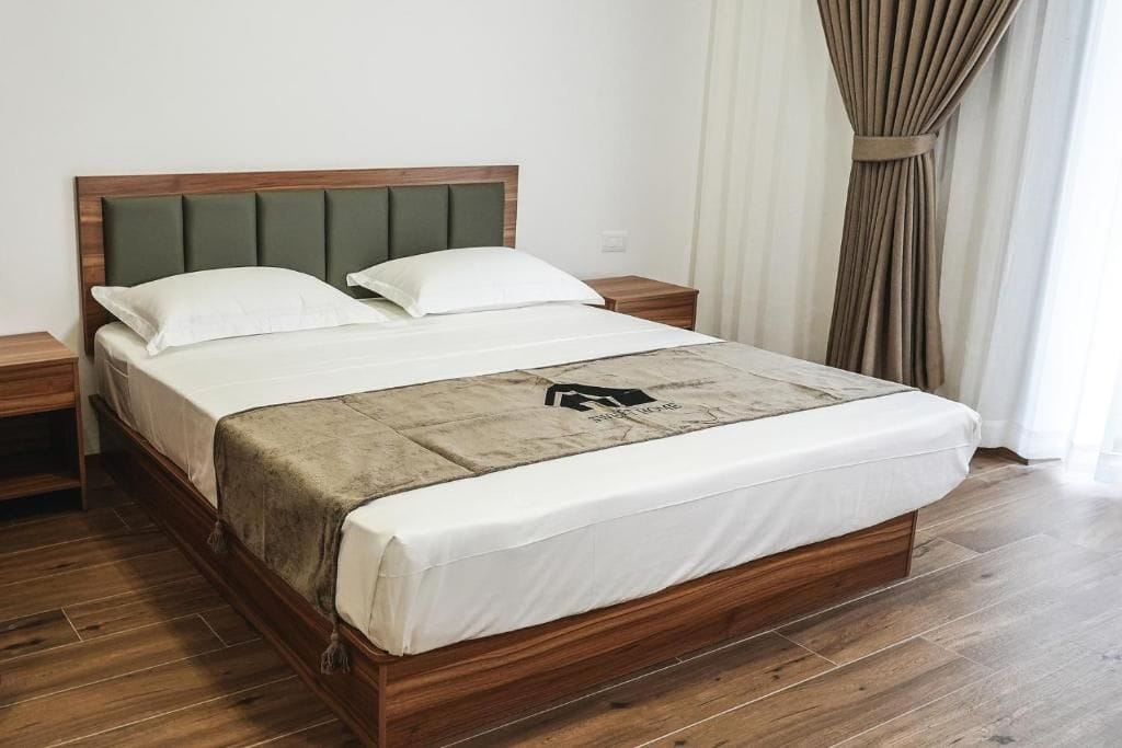 Una cama con estructura de madera, dos almohadas, una colcha blanca y un edredón con un logotipo.