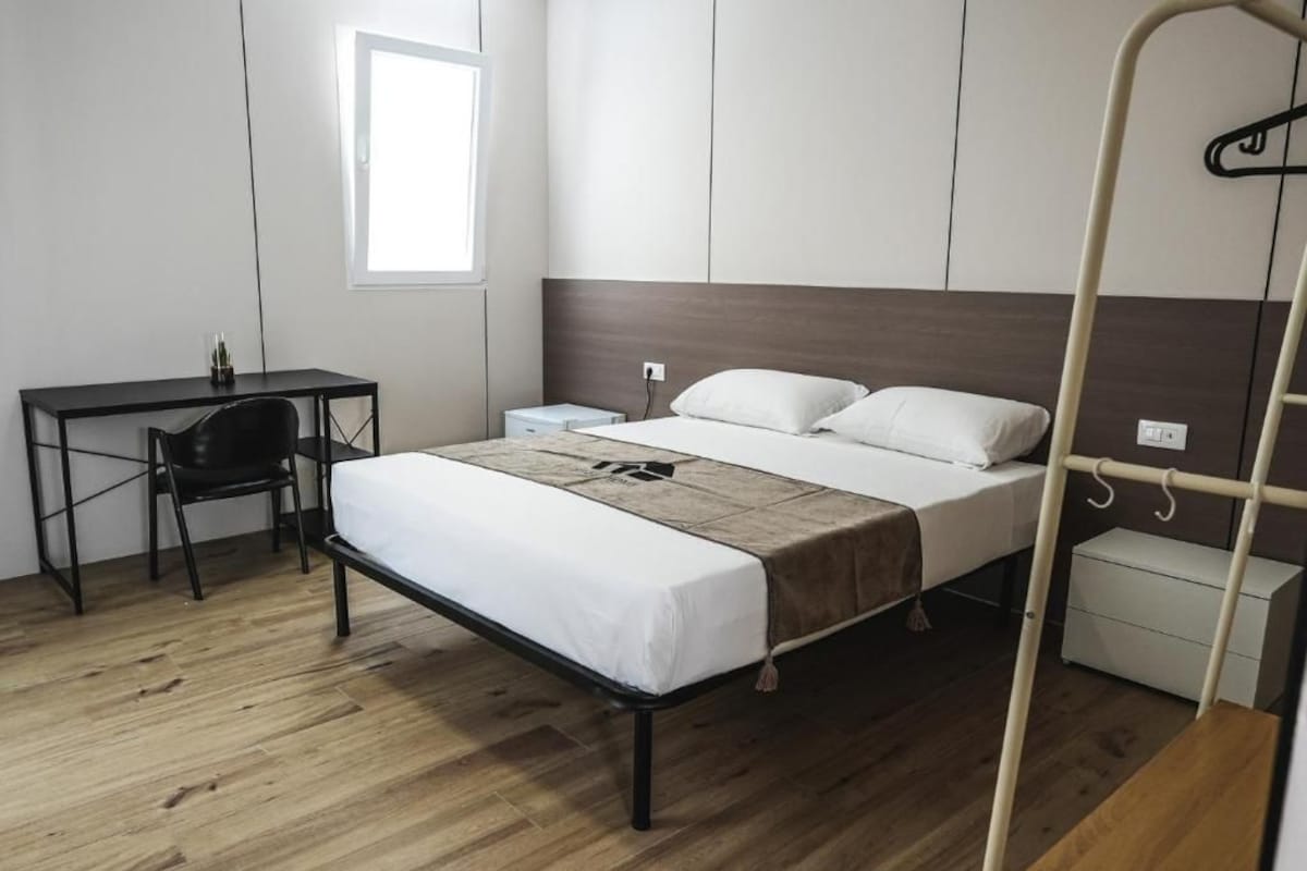Een slaapkamer met een bed, nachtkastje, stoel, bureau en een raam.