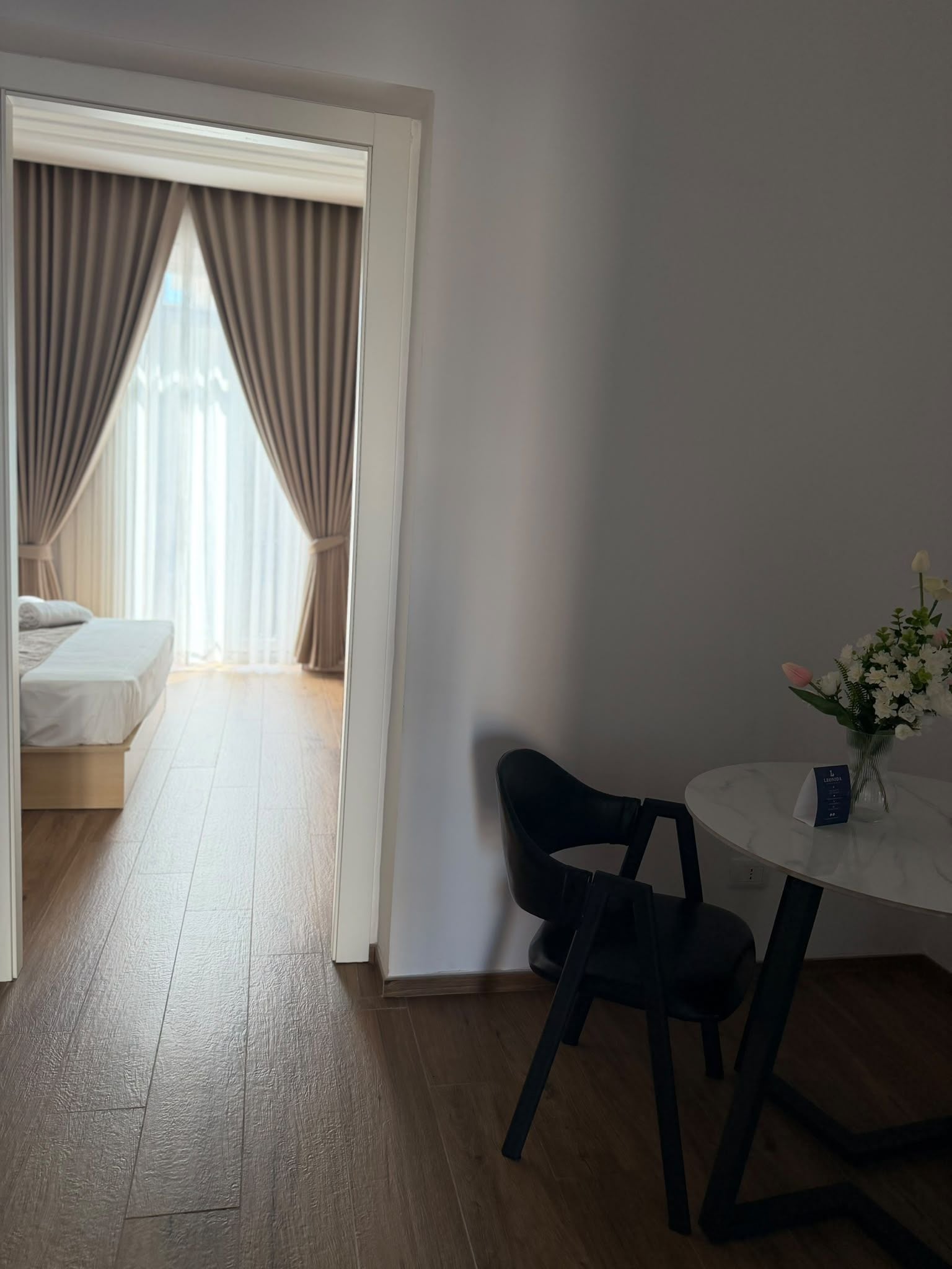 Une chambre d’hôtel avec un lit, un bureau et une chaise, un vase avec des fleurs et une vue sur l’extérieur à travers une fenêtre avec des rideaux.