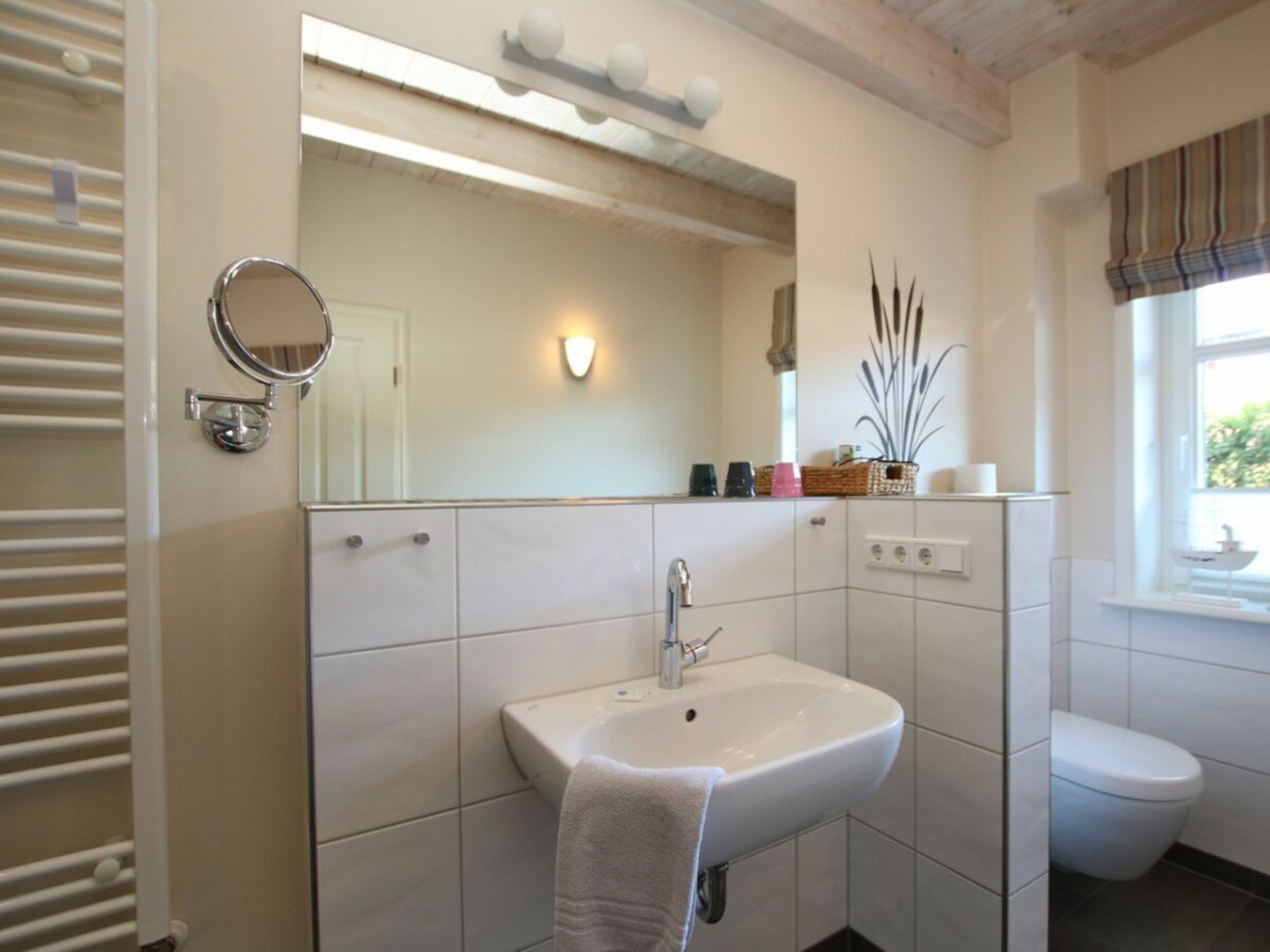 Een badkamer met een witte wastafel, spiegel en toilet. Een handdoek hangt aan de muur, en een raam met uitzicht op groen.