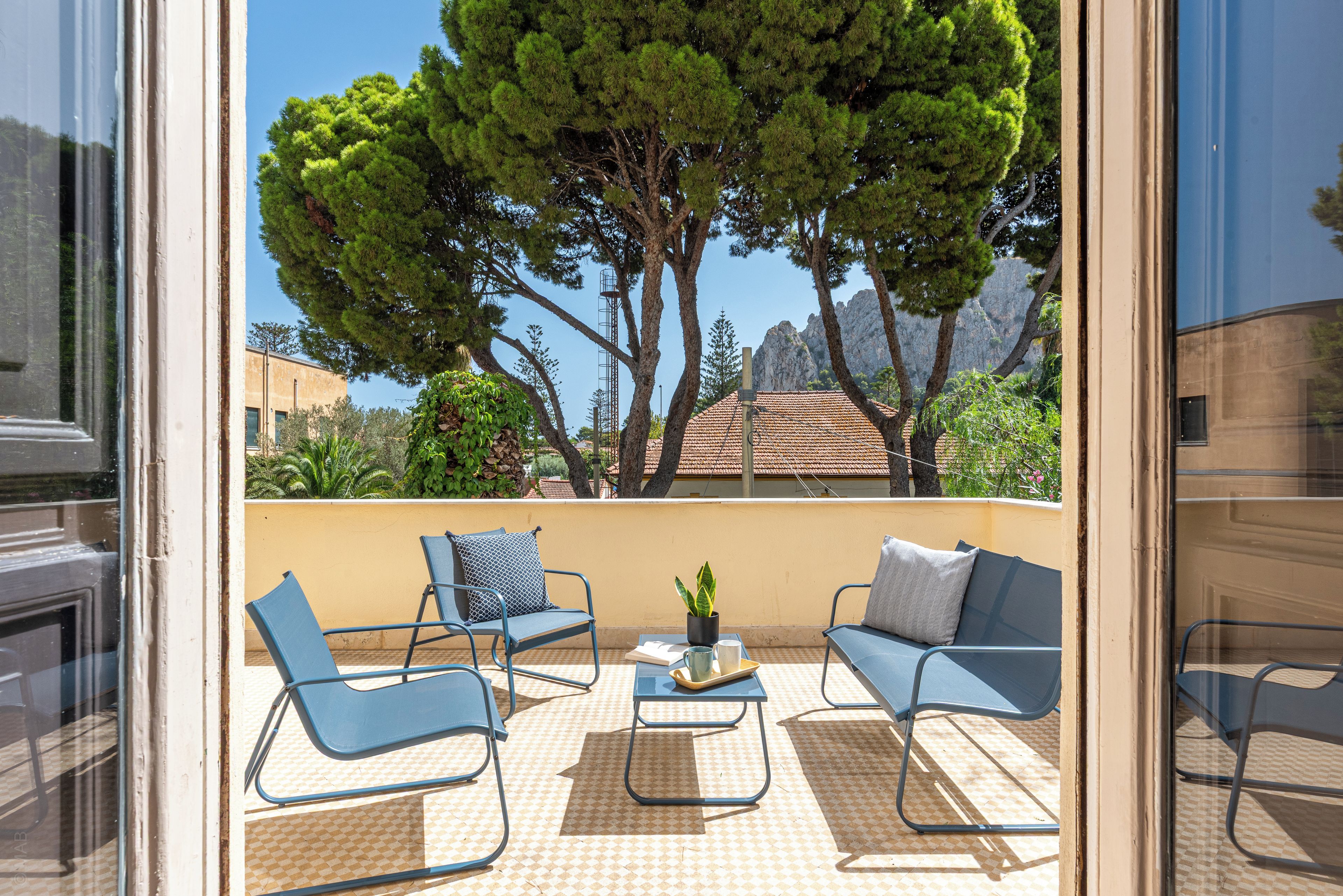 A balcony with two blue chairs, a small table with a plant, and a view of trees and buildings.