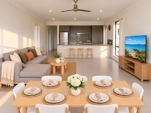 Una sala moderna con sofá, mesa de centro y mesa de comedor lista para seis comensales. Se ve una cocina con refrigerador y estufa al fondo.