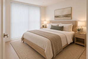 Une chambre à coucher avec un grand lit, deux tables de chevet avec des lampes, une fenêtre avec des stores et un tableau au mur.