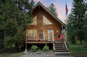 A log cabin with a porch, a flag, and a view of the surrounding forest. - Scenic Island Park Cabin with Private Hot Tub & ATV Access Near Yellowstone, Idaho (Island Park)