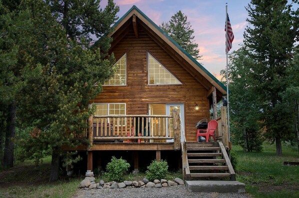 A log cabin with a porch, a flag, and a view of the surrounding forest. - Scenic Island Park Cabin with Private Hot Tub & ATV Access Near Yellowstone, Idaho (Island Park)