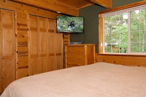 A bedroom with a bed, wooden wardrobe, a television mounted on the wall, and a window with a view of trees. - Scenic Island Park Cabin with Private Hot Tub & ATV Access Near Yellowstone, Idaho (Island Park)