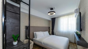 Habitación de hotel moderna con una cama grande, una mesita de noche, una planta en maceta, un televisor montado en la pared y una lámpara en el techo.