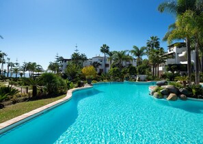 Una piscina blava i cristal·lina envoltada de palmeres i un edifici blanc amb balcons.