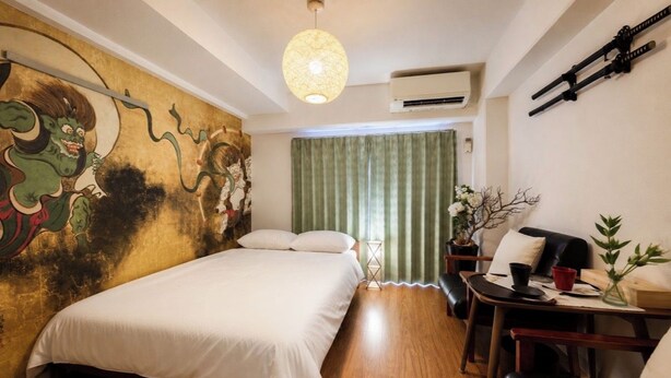 A hotel room with a bed, a desk with a chair, a wall-mounted air conditioner, and a decorative wall piece. - Vrbo Property (板橋区)