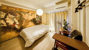 A hotel room with a bed, a desk, a chair, a wall with traditional artwork, and an air conditioning unit. - Vrbo Property (板橋区)