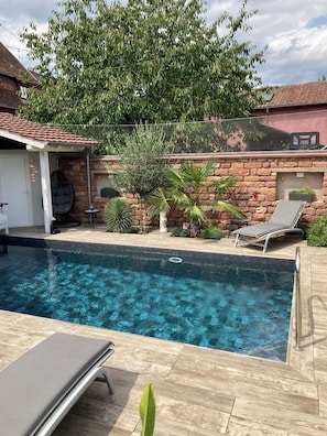 A swimming pool with a wooden deck, a stone wall, and a hanging chair. - Vrbo Property (Gertwiller)