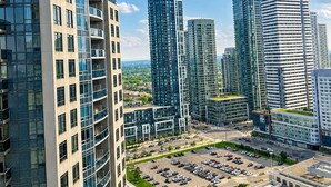 A high-rise building with a curved glass facade, surrounded by other modern high-rises, a parking lot, and a clear sky. - High-Rise 3BR Oasis | Free Parking | City Views (Mississauga)