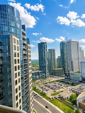 A high-rise building with a curved glass facade, surrounded by other modern high-rises, a parking lot, and a clear sky. - High-Rise 3BR Oasis | Free Parking | City Views (Mississauga)