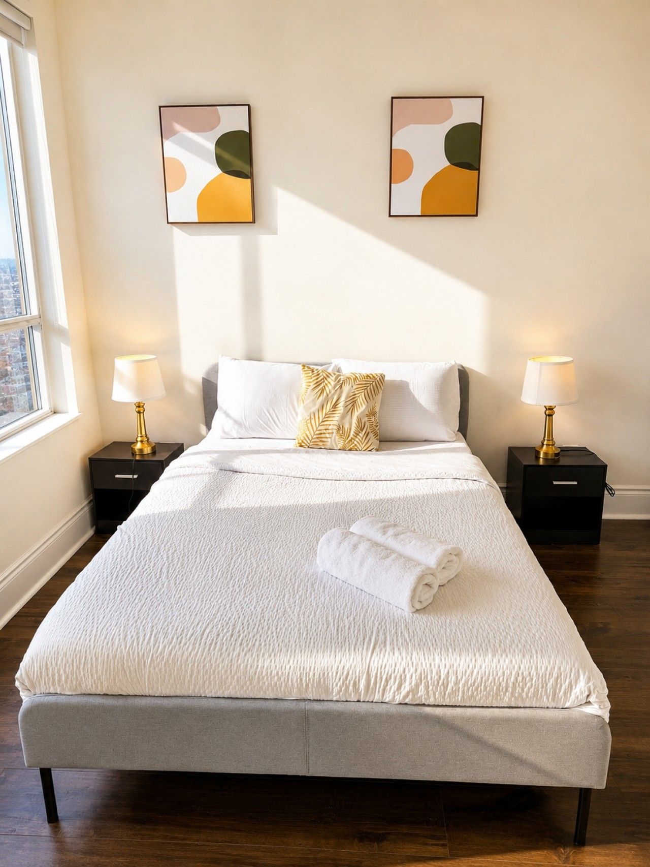 A neatly made bed with a white comforter and a rolled towel, two bedside tables with lamps, and abstract wall art.