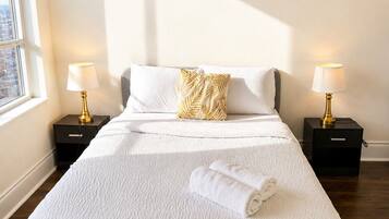 A neatly made bed with a white comforter and a rolled towel, two bedside tables with lamps, and abstract wall art.
