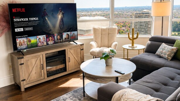 A modern living room with a large TV displaying a Netflix series, a sofa set, a coffee table, and a view of a cityscape through large windows. - High-Rise 3BR Oasis | Free Parking | City Views (Mississauga)