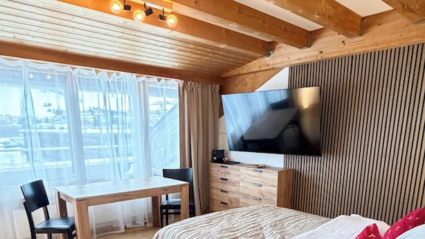 A modern bedroom with a wooden ceiling, a flat-screen TV mounted on the wall, a bed with a white comforter, a bedside table with a lamp, and a large window with sheer curtains. - Plan-B Stay (Samedan)