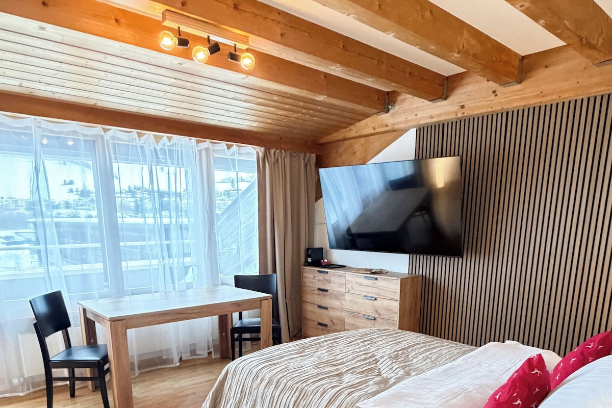 A modern bedroom with a wooden ceiling, a flat-screen TV mounted on the wall, a bed with a white comforter, a bedside table with a lamp, and a large window with sheer curtains.