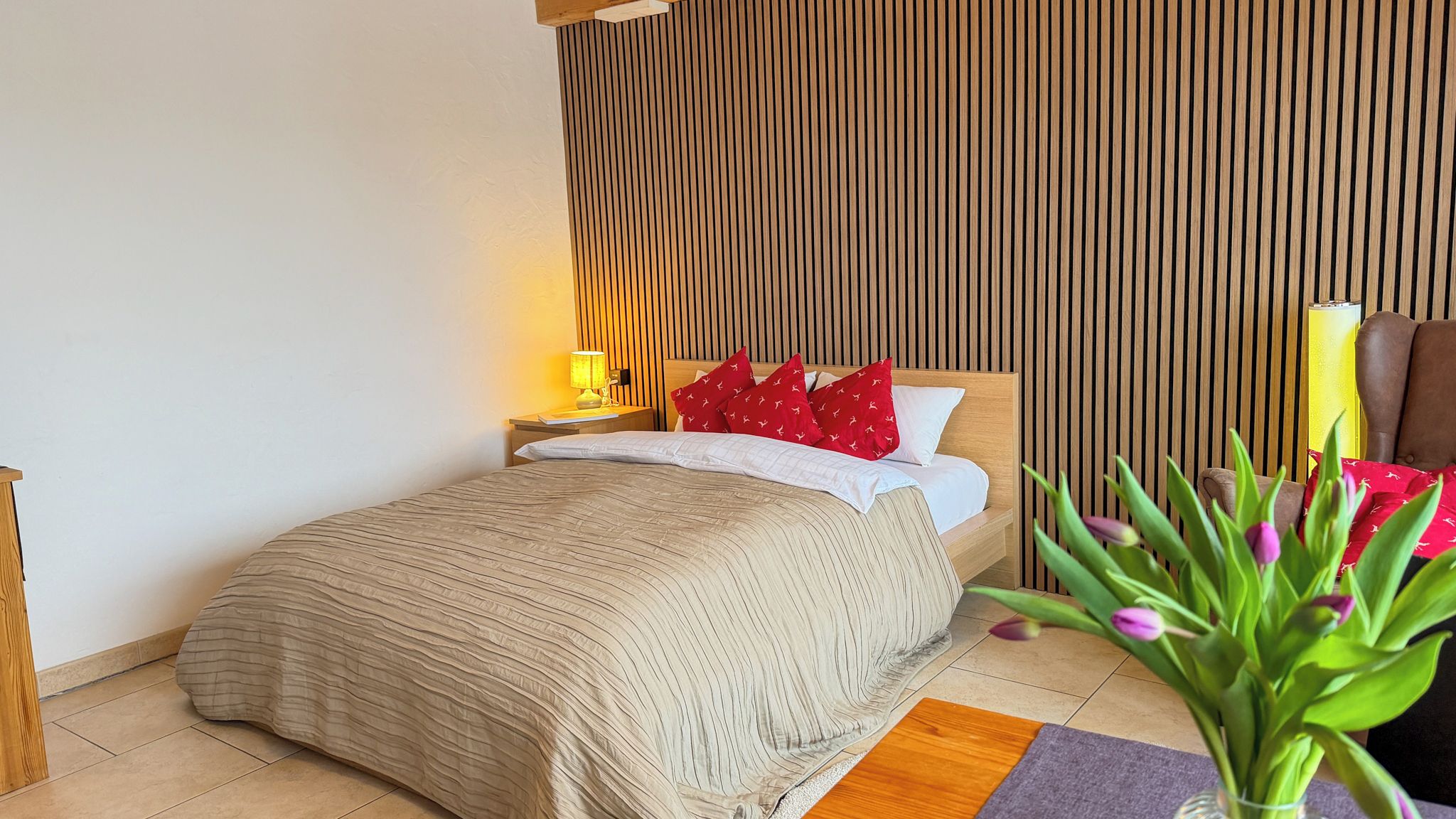 A bedroom with a bed, bedside tables, a lamp, a chair, and a vase with flowers.