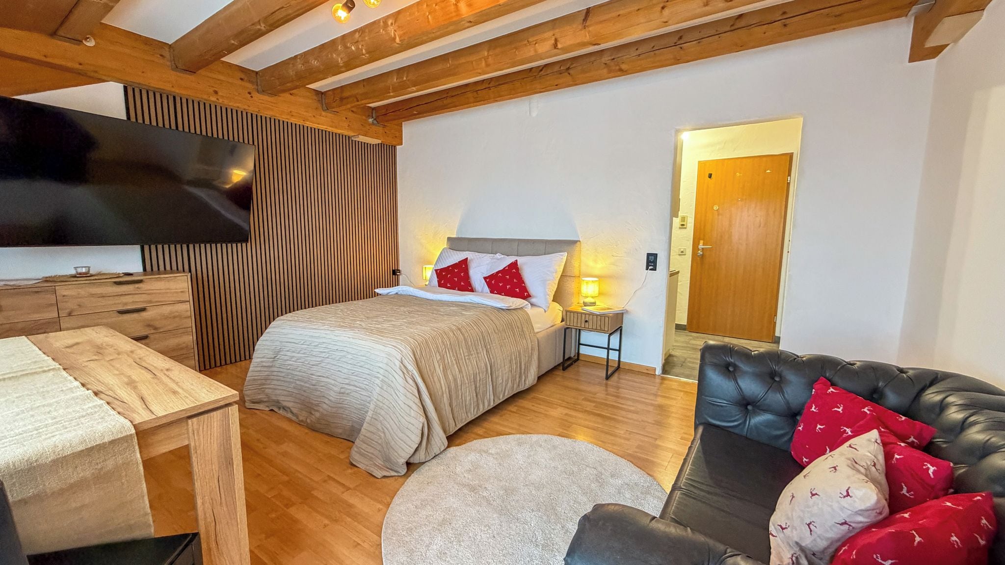 Un dormitorio con una cama, mesitas de noche, un televisor, un aparador de madera, un sofá de cuero y una puerta que da a otra habitación.